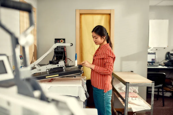Experto en impresión. Joven trabajadora alegre utilizando la máquina de transferencia de prensa de calor para imprimir pegatina publicitaria con texto en la camiseta en el lugar de trabajo — Foto de Stock