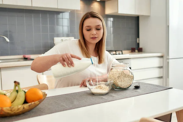 Wo die Haferflocken zuerst kommen. Junge Frau gießt Milch in eine Schüssel mit Hafer auf den Tisch, während sie in der Küche das Frühstück zubereitet — Stockfoto