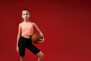 Oynamaya hazırız. Tatlı bir çocuk, kız sporla meşgul, basketbol oynarken kameraya bakıyor. Kırmızı arka planda izole edilmiş. Fitness, eğitim, aktif yaşam tarzı konsepti. Yatay çekim