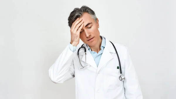 Tıbbi üniformalı yorgun ve endişeli bir doktor. Steteskopla boynuna ve alnına dokunurken gri arka planda duruyor. — Stok fotoğraf
