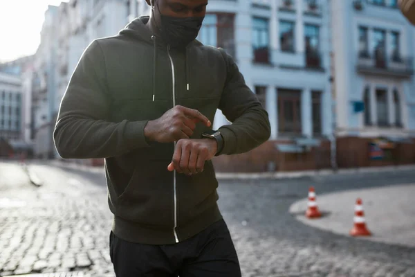 Getting ready to run. Young african male runner in black sportswear and medical protective mask touching his smartwatch while standing at the empty street