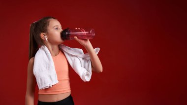 Sıvı al. Şirin bir kız sporla meşgul, havluyla ve içme suyuyla dikilirken yana bakıyor. Kırmızı arka planda izole edilmiş. Fitness, eğitim, aktif yaşam tarzı konsepti. Yatay çekim