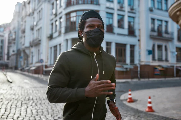 Running During Quarantine. Young african athletic man in black sportswear and medical protective mask running through the city street in the morning
