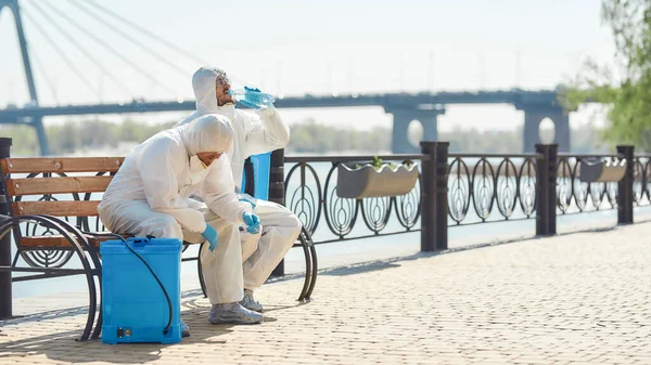 Exhausto. Saneamiento, limpieza y desinfección de la ciudad debido a la aparición del virus Covid19. Equipo especializado en trajes de protección y máscaras se toma un descanso cerca de la orilla del río — Foto de Stock