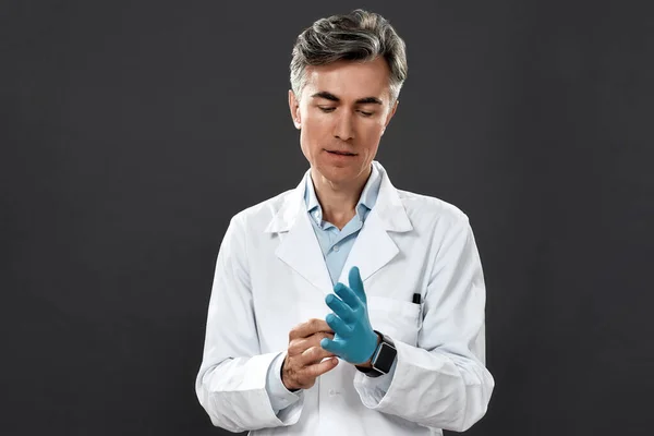 Egy orvosegyenruhás, érett férfi orvos portréja, aki védőruhát visel, steril kék kesztyűt, miközben fekete háttér előtt áll. — Stock Fotó