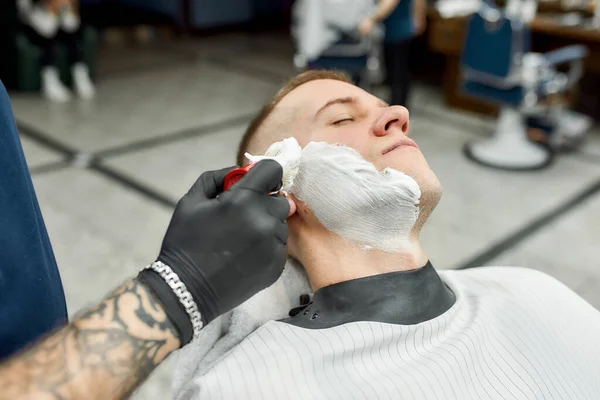 Correção de barba. Barbeiro em luvas pretas com tatuagem no braço aplicando espuma de barbear no rosto de um jovem sentado em uma cadeira de barbearia. Limpeza e barbear — Fotografia de Stock