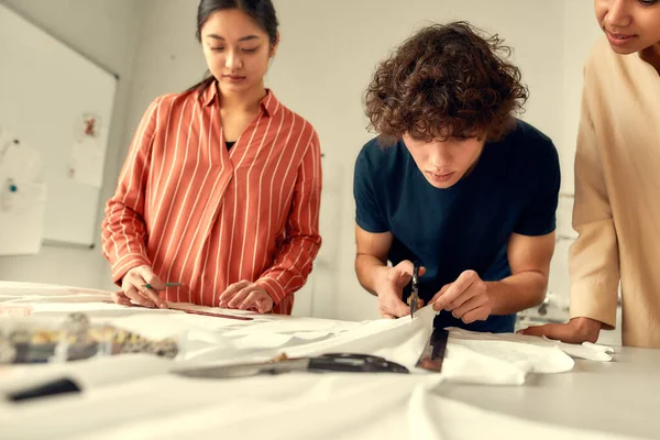 Zvýšit kvalitu krejčovství. Mladý návrhář stříhající bílou textilii ve studiu. Skupina kreativních milénií spolupracujících — Stock fotografie