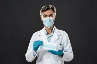 Kendine dikkat et. Tıbbi üniformalı ve mavi steril eldivenli olgun bir erkek doktor siyah arka planda dururken kameraya tıbbi maske takıyor.