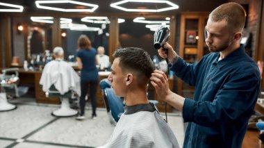Modern berber dükkanını ziyaret ediyorum. Erkek berberin müşterilerinin saçlarını kurutuşunun yan görüntüsü. Berber dükkanında yeni saç kesimi yaptırıyorum. Seçici görünüm