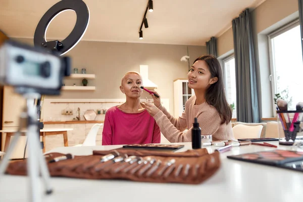 Tudo por ser uma rapariga. Blogger feminino asiático aplicando maquiagem no rosto de mulher de meia-idade usando produtos cosméticos na mesa. Duas mulheres gravando um vídeo tutorial para blog de beleza — Fotografia de Stock