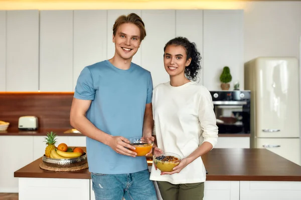 Φαγητό που σε κάνει να νιώθεις καλά. Ευτυχισμένο ζευγάρι χαμογελά στην κάμερα, ενώ στέκεται στην κουζίνα μαζί. Νεαρός άνδρας και γυναίκα κρατώντας μπολ smoothie. Χορτοφαγία, υγιεινά τρόφιμα, διατροφή, μείνετε στο σπίτι έννοια — Φωτογραφία Αρχείου