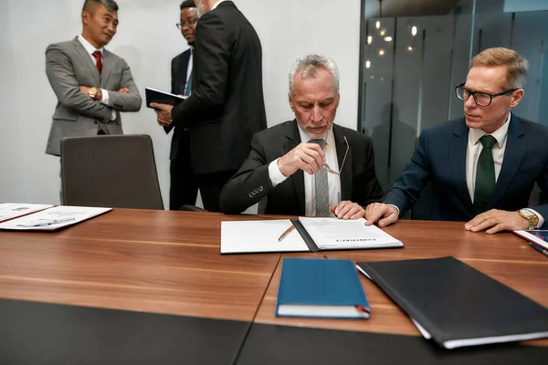 Tengo que pensarlo. Hombre de negocios maduro y seguro con gafas y leyendo un contrato mientras tiene una reunión con sus socios de negocios en la oficina moderna — Foto de Stock