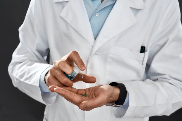 Disinfecting hands. Close up photo of a male doctor in medical uniform using sanitizer or antiseptic gel while standing against black background