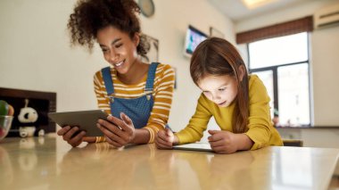 Bebek için harika bir zaman. Afro-Amerikalı kadın bebek bakıcısı ve tablet bilgisayar kullanan beyaz bir kız evde oturuyor. Çocuk eğitimi, boş zaman aktiviteleri, bebek bakıcılığı konsepti