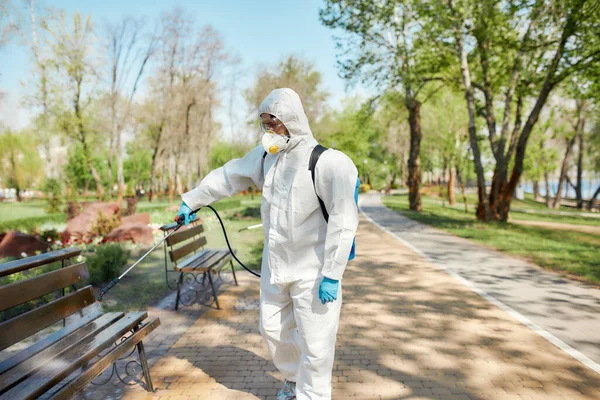 Υπηρεσία που μπορείς να εμπιστευτείς. Εξυγιάνσεις, καθαρισμός και απολύμανση του πάρκου της πόλης λόγω της εμφάνισης του ιού Covid19. Εργαζόμενος με προστατευτική στολή και μάσκα — Φωτογραφία Αρχείου
