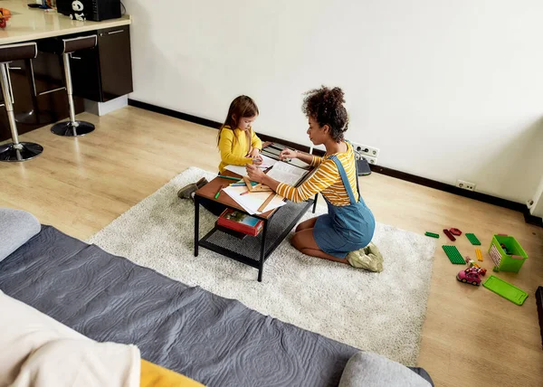 Menina caucasiana passando tempo com babá afro-americana. Eles estão desenhando, aprendendo a escrever cartas, sentados no chão — Fotografia de Stock