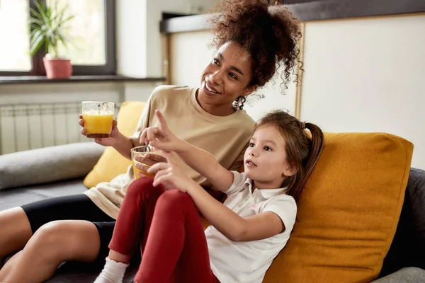 Spending time together. African american woman baby sitter entertaining caucasian cute little girl. They are sitting on the couch, drinking juice while watching TV — Stock Photo, Image