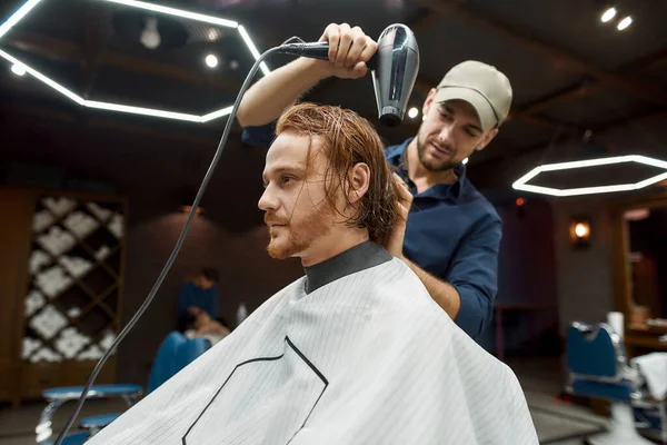 Вид сбоку на молодого профессора-парикмахера, сушащего волосы красивого рыжего парня, сидящего в парикмахерском кресле перед зеркалом в современной парикмахерской — стоковое фото