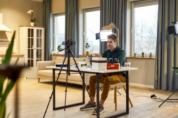 Traer alegría. Joven alegre blogger de tecnología masculina sonriendo, mientras graba video blog o vlog sobre nuevas lentes de cámara y otros gadgets sentados en el estudio en casa. Blogging, Trabajo desde el hogar concepto —  Fotos de Stock