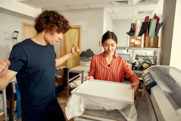 İstediğin her stili sağlıyorsun. Genç işçiler, erkek ve kadın işyerindeki ipek ekran baskı makinesinde baskı yapmak için tişört hazırlıyorlar. — Stok fotoğraf