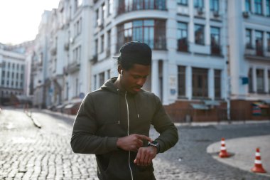 Sabah koşusu. Siyah spor giysili Afrikalı genç bir adam spor ayakkabısına dokunuyor, spor uygulamasını boş sokakta dururken kullanıyor.