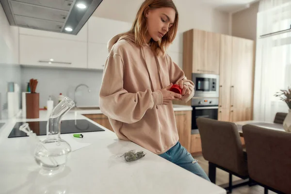 Préparation. Jeune femme utilisant un broyeur à marijuana rouge, debout dans la cuisine. Tuyau d'eau en verre ou bong, bourgeons de cannabis dans un sac en plastique sur la table. Concept de légalisation du cannabis et des mauvaises herbes — Photo