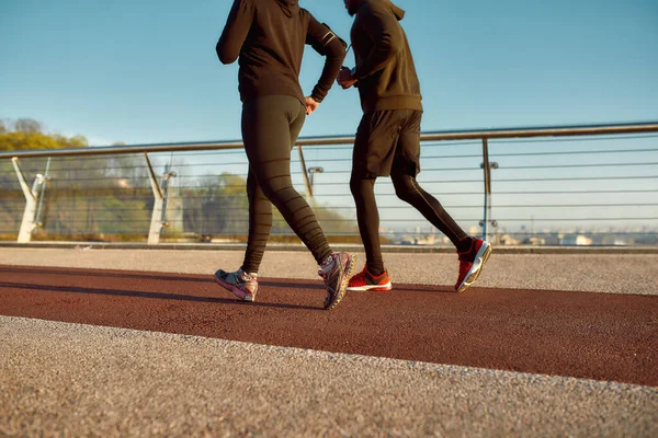 Pessoas a correr. Vista cortada de casal desportivo em sportswear preto correndo juntos na ponte pela manhã. Homem e mulher correndo ao ar livre — Fotografia de Stock