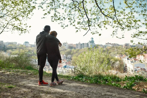 Largura completa de disparo de joven pareja de fitness en ropa deportiva mirando la increíble vista de la ciudad después de entrenar juntos al aire libre —  Fotos de Stock