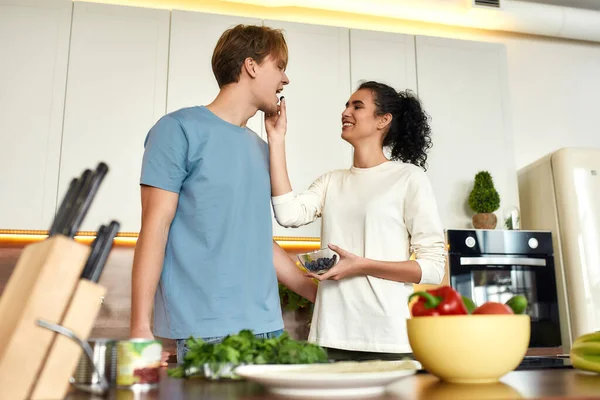 Šťastný pár, vegetariáni připravující zdravé jídlo, písek, salát v kuchyni spolu. Dívka krmí svého přítele borůvkami. Vegetariánství, zdravé jídlo, koncepce stravy — Stock fotografie