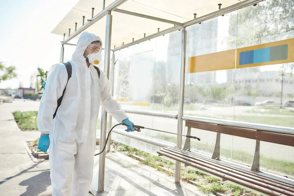 Now it is clean. Sanitization, cleaning and disinfection of the streets and alleys in the city due to the emergence of the Covid19 virus. Man in protective suit and mask at work near the bus stop — Stock Photo, Image