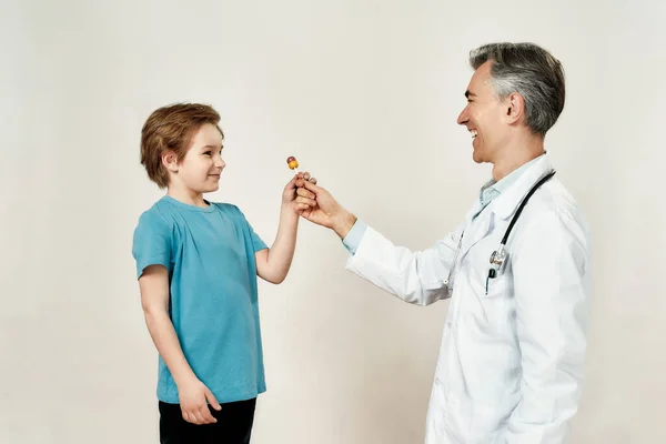 Después del chequeo exitoso. Amable pediatra masculino en uniforme médico dando una piruleta para niño feliz mientras está de pie sobre fondo gris — Foto de Stock