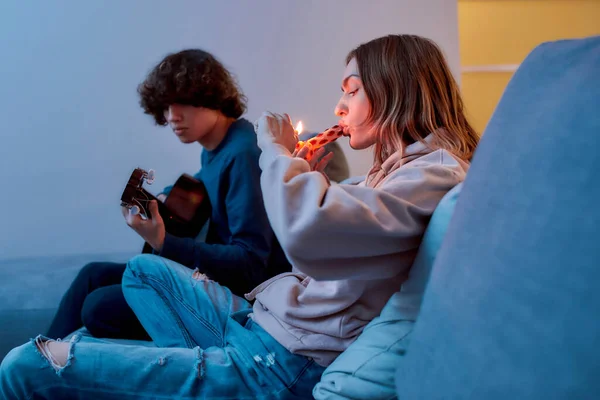 Accesorios para fumar. Niña encendiendo cannabis en pipa de cristal o pipa, relajándose con su novio en el sofá en casa. Chico tocando en la guitarra — Foto de Stock