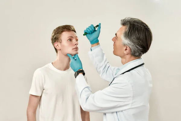 Ophthalmology clinic. Professional mature male ophthalmologist in medical uniform examining eyes of young patient while standing against grey background