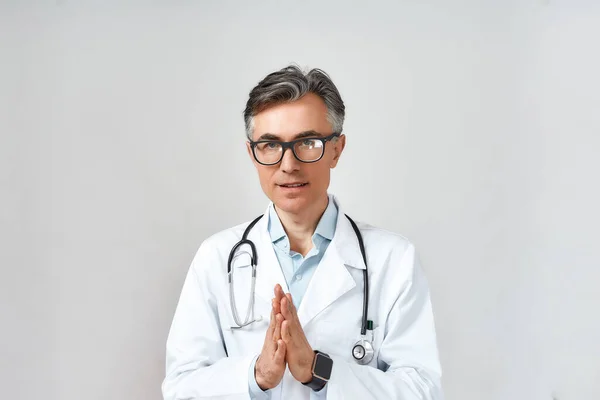 Portré érett férfi orvos vagy orvos fehér köpenyben sztetoszkóppal a nyak körül imádkozik Istenhez segítségért, miközben áll szemben szürke háttér — Stock Fotó