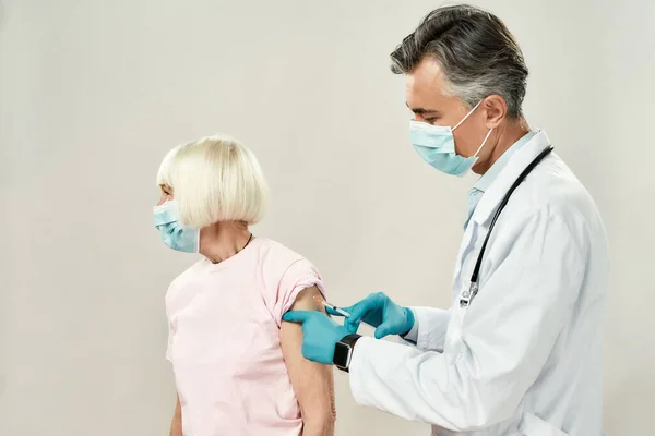 Vaccines for the elderly. Professional male doctor in blue sterile gloves injecting vaccine to senior woman wearing medical mask. Vaccination, medicine and healthcare