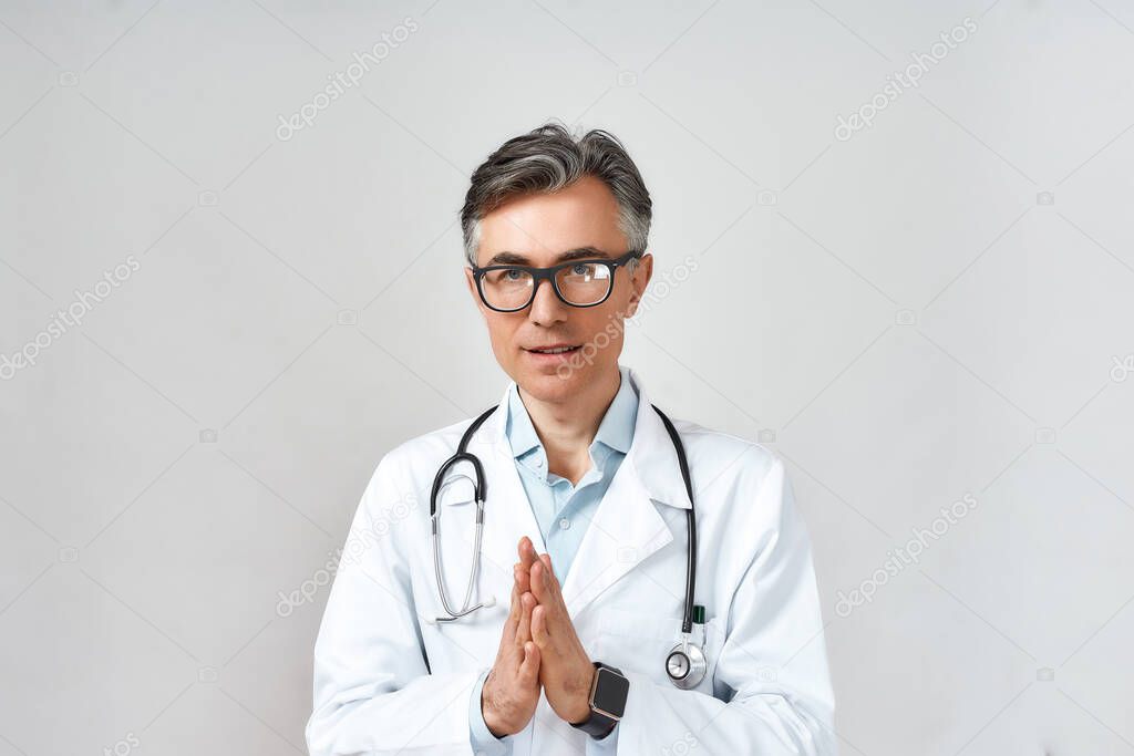 Portrait of mature male doctor or physician in white coat with stethoscope around neck praying to God for help while standing against grey background