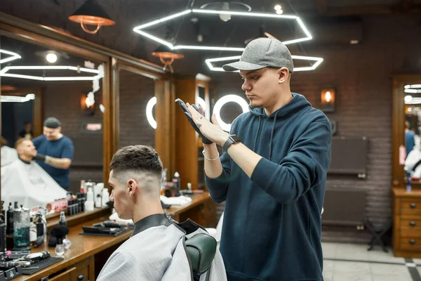 Arbeiten im Friseursalon. Professioneller Friseur mit Baseballmütze frisiert junge und gutaussehende Kerle, die im Stuhl vor einem Spiegel sitzen — Stockfoto