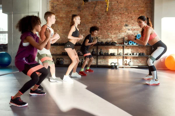 Colpo completo di bambini che si allenano usando una fascia di resistenza in palestra insieme all'allenatore donna. Sport, stile di vita sano, educazione fisica — Foto Stock