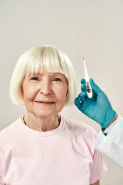 İyi hissediyorum. Mavi steril eldivenli doktor kafasının yakınındaki sıcaklığı ölçmek için elektronik termometre tutarken kameraya gülümseyen yaşlı kadının portresi.