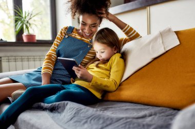 Kaliteli çocuk bakımı önemlidir. Afro-Amerikalı kadın bebek bakıcısı ve beyaz tatlı küçük kız gülümsüyor, tablet bilgisayar kullanıyor, evde oturuyor. Çocuk eğitimi, boş zaman aktiviteleri, bebek bakıcılığı konsepti