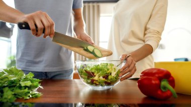 Kadın ona yardım ederken adamın dilimlenmiş salatalığı bir tabağa koyup salata tabağını tutarken görüntüsü. Vejetaryenler mutfakta birlikte sağlıklı bir yemek hazırlıyorlar.