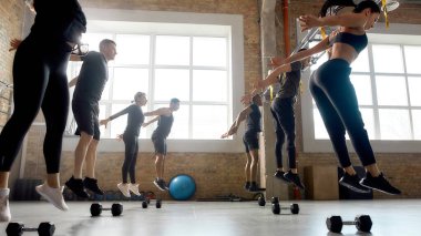 Cesetler yere düşsün. Bir grup sportif insan atlıyor, endüstriyel spor salonunda dambıllarla antrenman yapmadan önce ısınıyorlar.