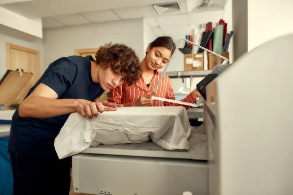 Ponte lo que quieras. Trabajadores jóvenes, hombres y mujeres están preparando la camiseta para imprimir en la máquina de serigrafía en el lugar de trabajo — Foto de Stock