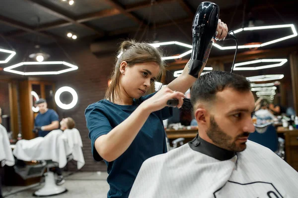 Getting new haircut. Young bearded handsome man sitting in barber shop chair while female barber drying his hair — Stock Photo, Image