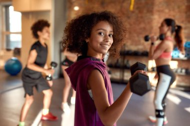 Spor salonunda halterle egzersiz yaparken kameraya gülümseyen Afrikalı Amerikalı kız, kadın antrenör ve diğer çocuklarla birlikte. Spor, sağlıklı yaşam tarzı, beden eğitimi konsepti
