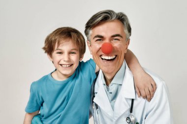 Sevimli, heyecanlı, palyaço burunlu neşeli erkek çocuk doktoruyla kucaklaşan küçük çocuğun portresi. Kameraya bakıp gülümsüyorlar. Çocuklarla çalışan doktor.