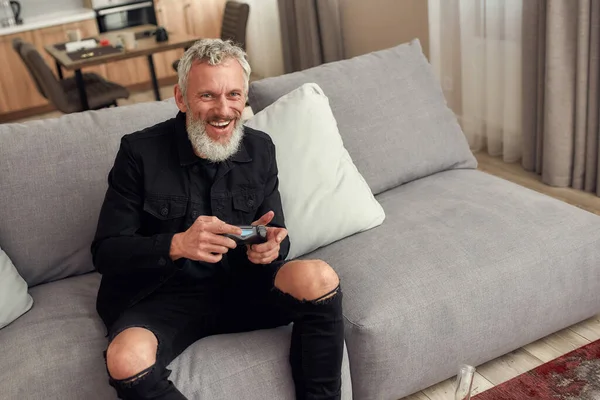 Blijf aan de top. Man van middelbare leeftijd met baard, die videospelletjes speelt nadat hij marihuana heeft gerookt uit een waterpijp of een glazen waterpijp die thuis op de bank zit — Stockfoto