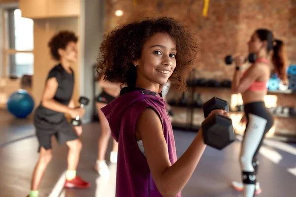 Menina afro-americana sorrindo para a câmera enquanto se exercita usando haltere no ginásio, juntamente com o treinador feminino e outras crianças. Esporte, estilo de vida saudável, conceito de educação física — Fotografia de Stock