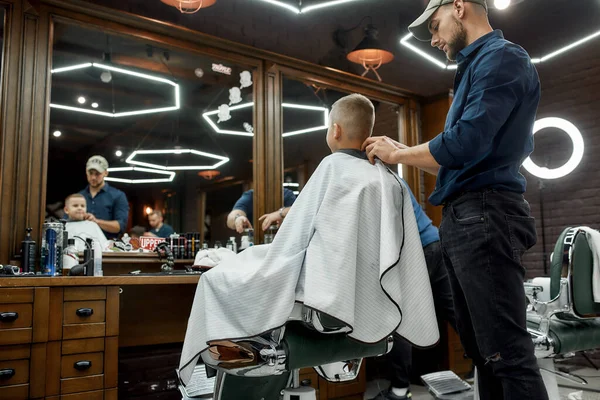 Great job. Young barber and little boy in barbershop. Back view. Hairdresser is taking off hairdresser cape