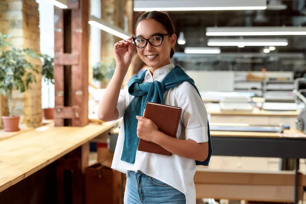 Genç ve başarılı. Gündelik kıyafetlerle gezen Asyalı çekici iş kadınının portresi dizüstü bilgisayarını tutuyor ve modern ofisteyken gözlüklerini ayarlıyor. — Stok fotoğraf